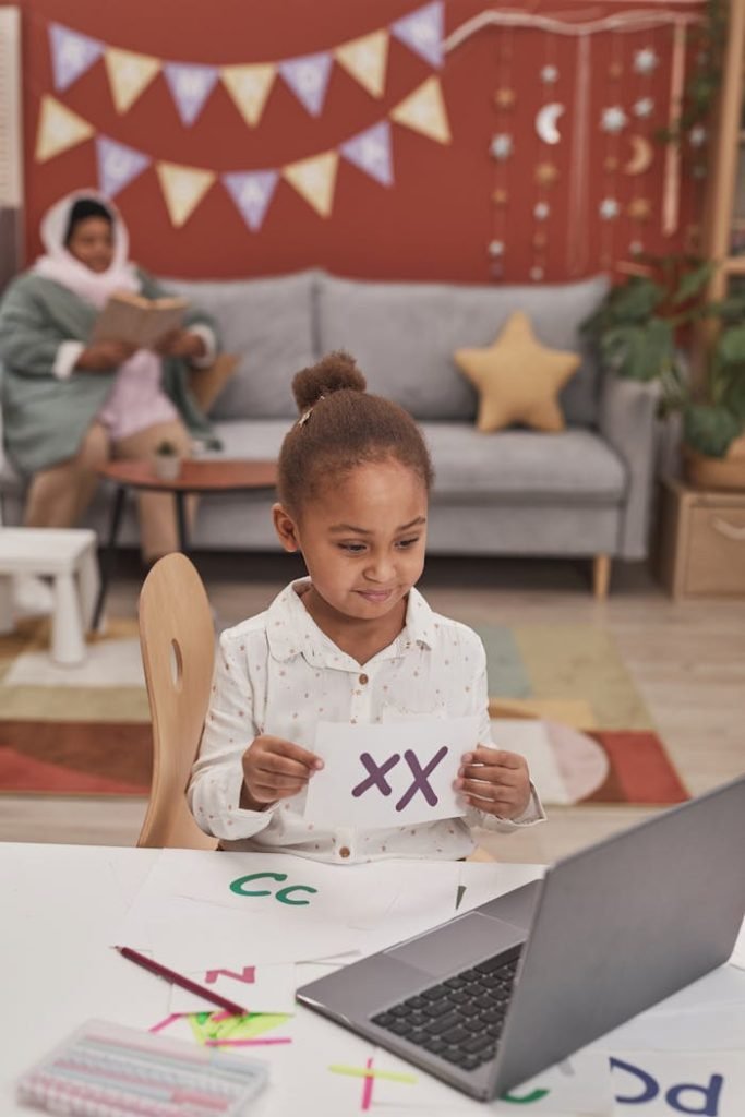 Child practicing alphabet through online learning with laptop at home. Mother watches in background.