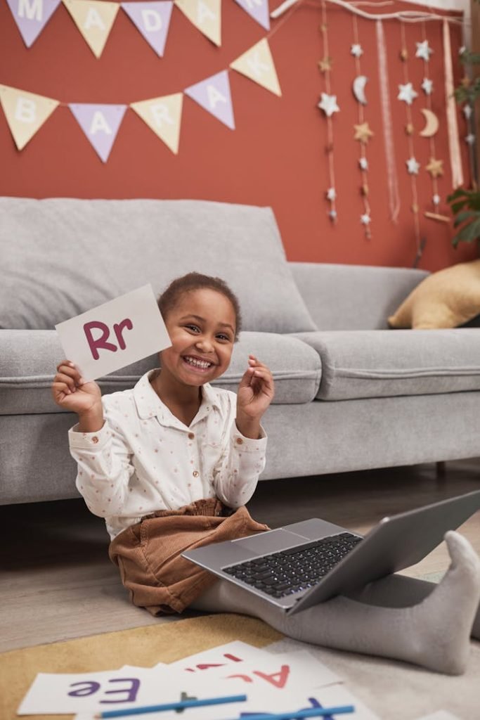 Smiling child using a laptop for online learning, holding a letter card indoors.